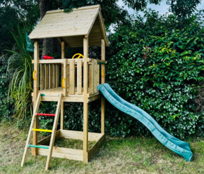 Kompaktes Holzspielhaus mit grüner Rutsche und Kletterleiter neben einer Hecke